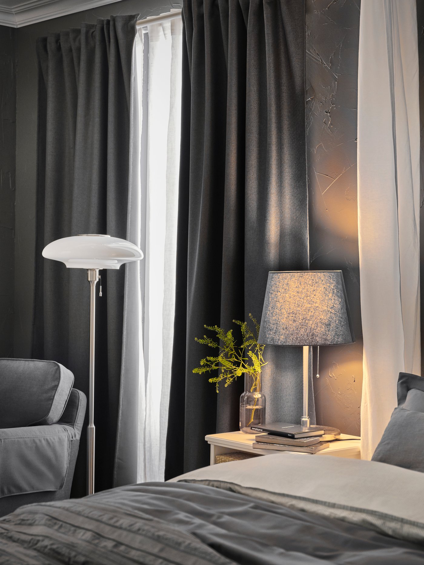 A bed with grey bed linen, a white bedside table with a grey table lamp and a white floor lamp, beside grey curtains.