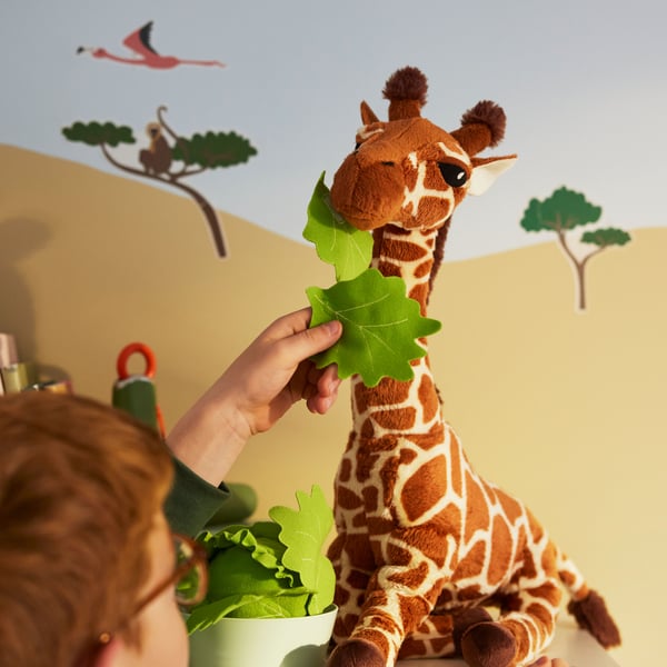 A child holds up a green leaf to feed a SANDLÖPARE soft toy giraffe.
