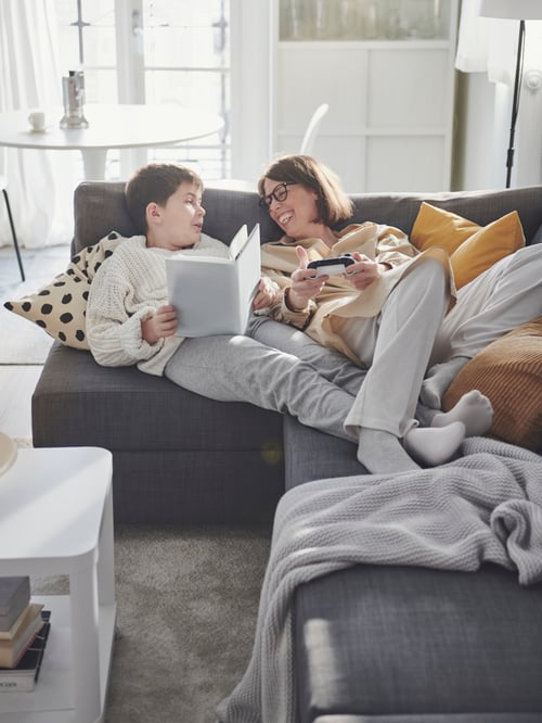 Eine Erwachsene und ein Kind liegen entspannt auf einem FRIHETEN Eckbettsofa mit Aufbewahrung und lesen. Neben ihnen steht ein TINGBY Beistelltisch mit Rollen.