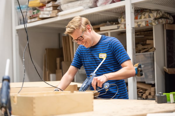IKEA medewerker in een blauwgestreept IKEA uniform glimlacht terwijl hij werkt met een elektrisch lijmpistool in een werkplaats.