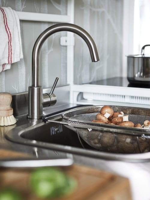 A GLYPEN stainless steel mixer tap on a sink holding a sieve full of mushrooms. A brush and tea towel are behind the tap.