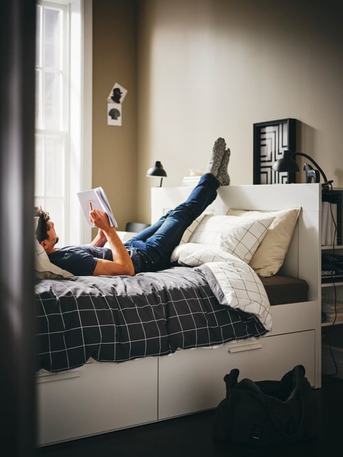 Bedroom area featuring a person reading on a white bed frame with storage and headboard, a duvet set, and a clamp spotlight.
