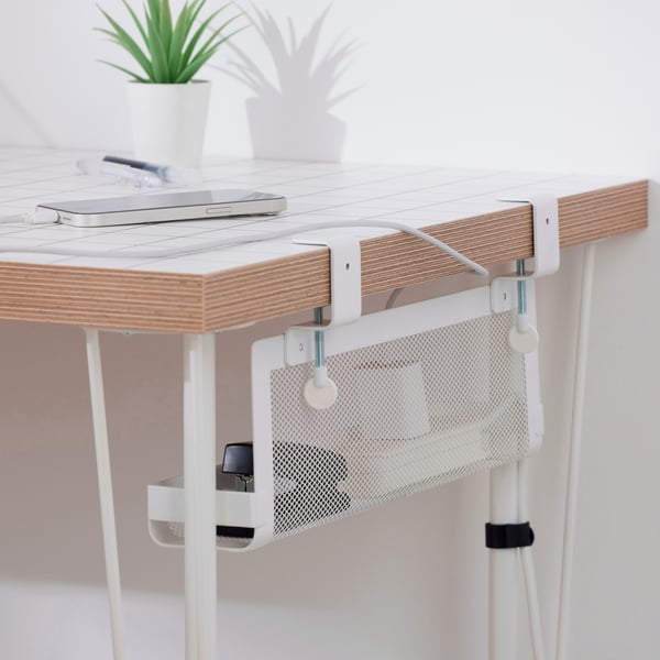 A white, steel mesh FÖRSÄSONG cable management tray is clamped onto and under the edge of a desk. It holds a power strip.