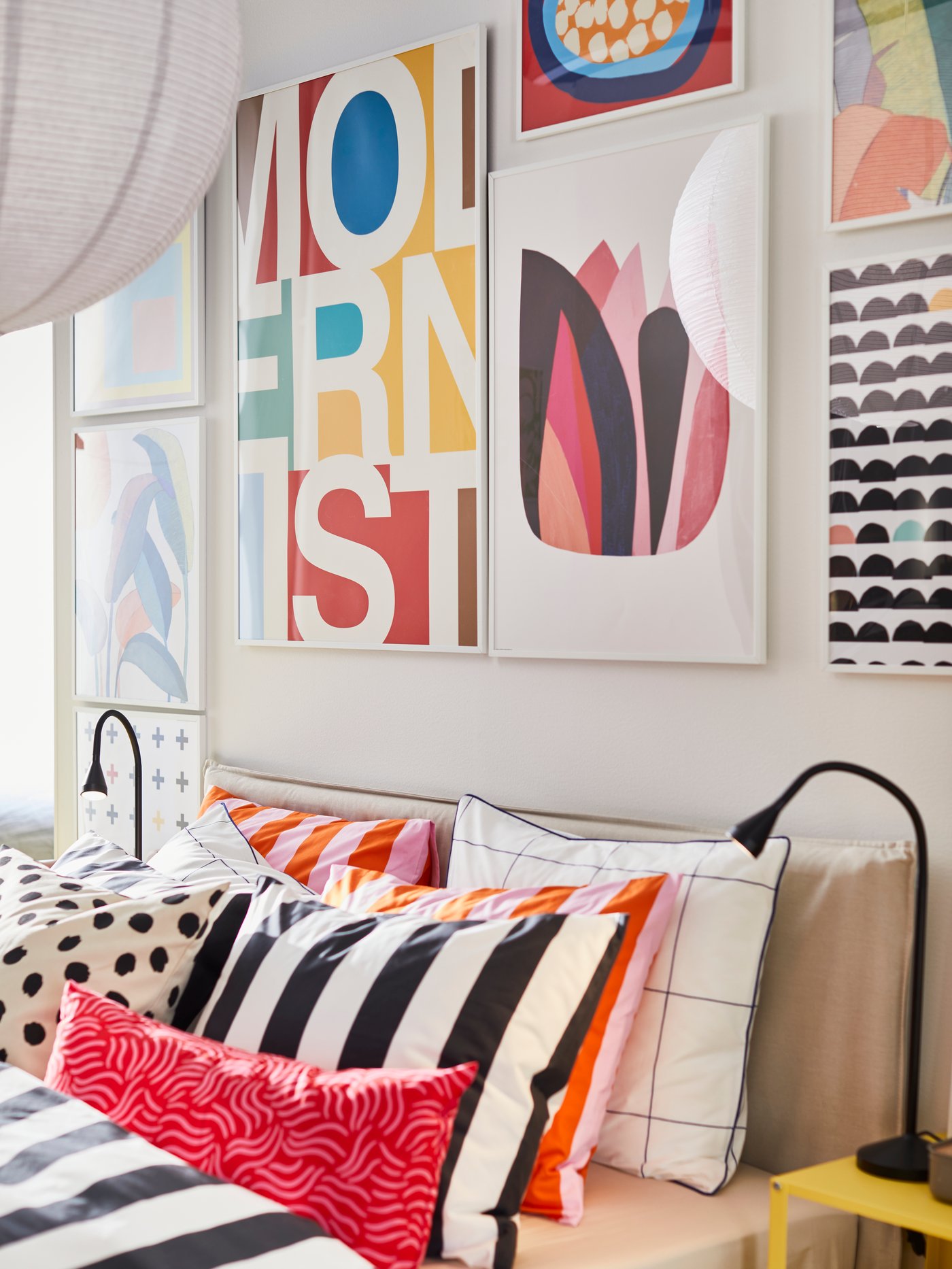A bed with colourful bedding has a gallery wall of modern artwork above it, including matching white RÖDALM frames.