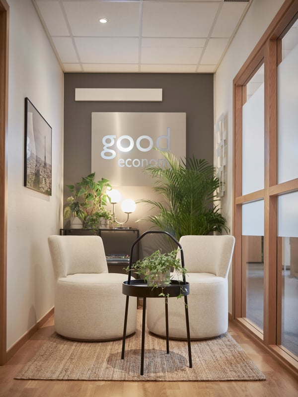 Litet rum på kontor med två vita fåtöljer mittemot varandra, runt svart bord med krukväxt, gröna växter runtomkring, upplyst väggskylt med texten 'good economy' och glasfönster från golv till tak till höger.