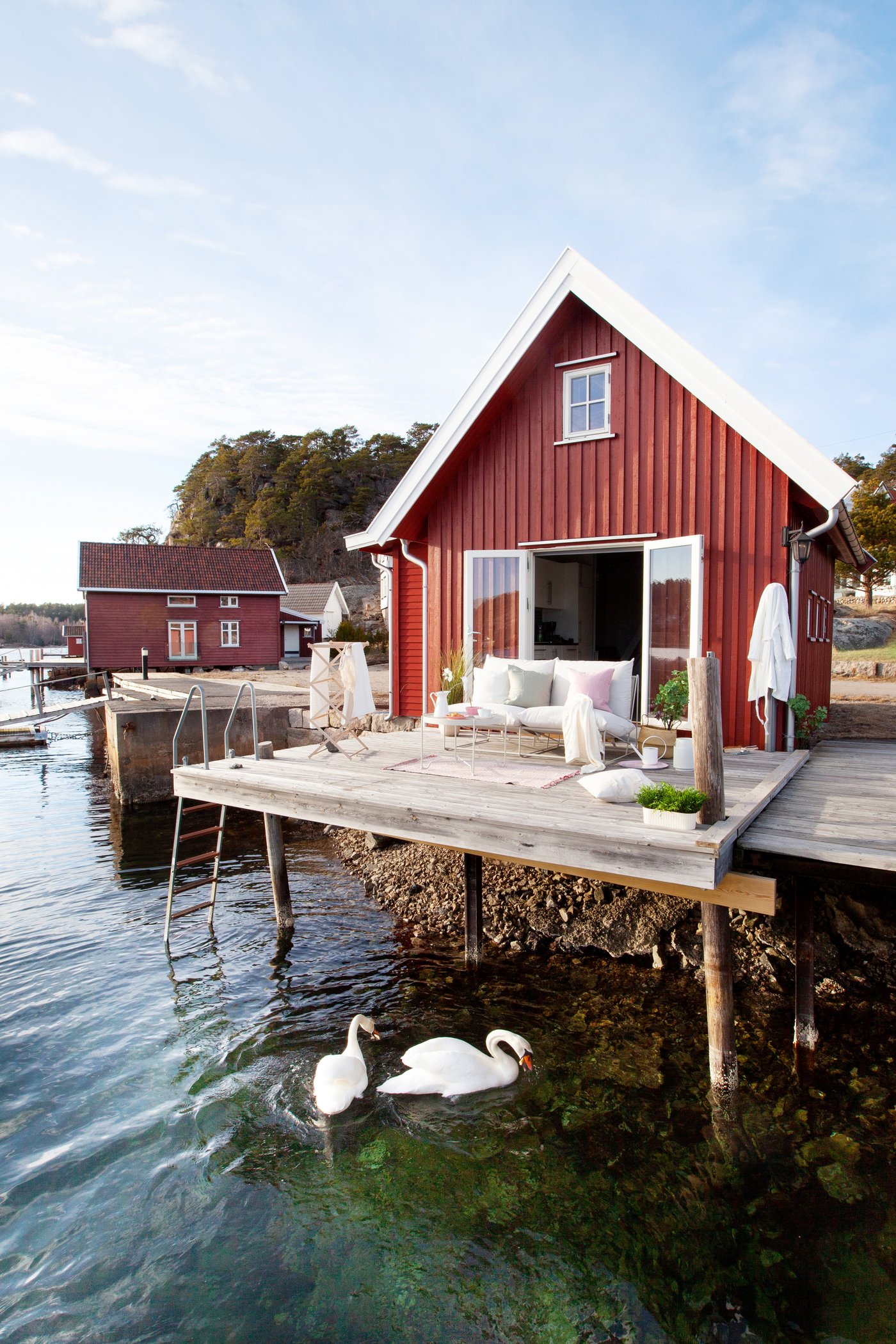 Et rødt sommerhus med en træterrase over vandet hvor 2 svaner svømmer rundt.