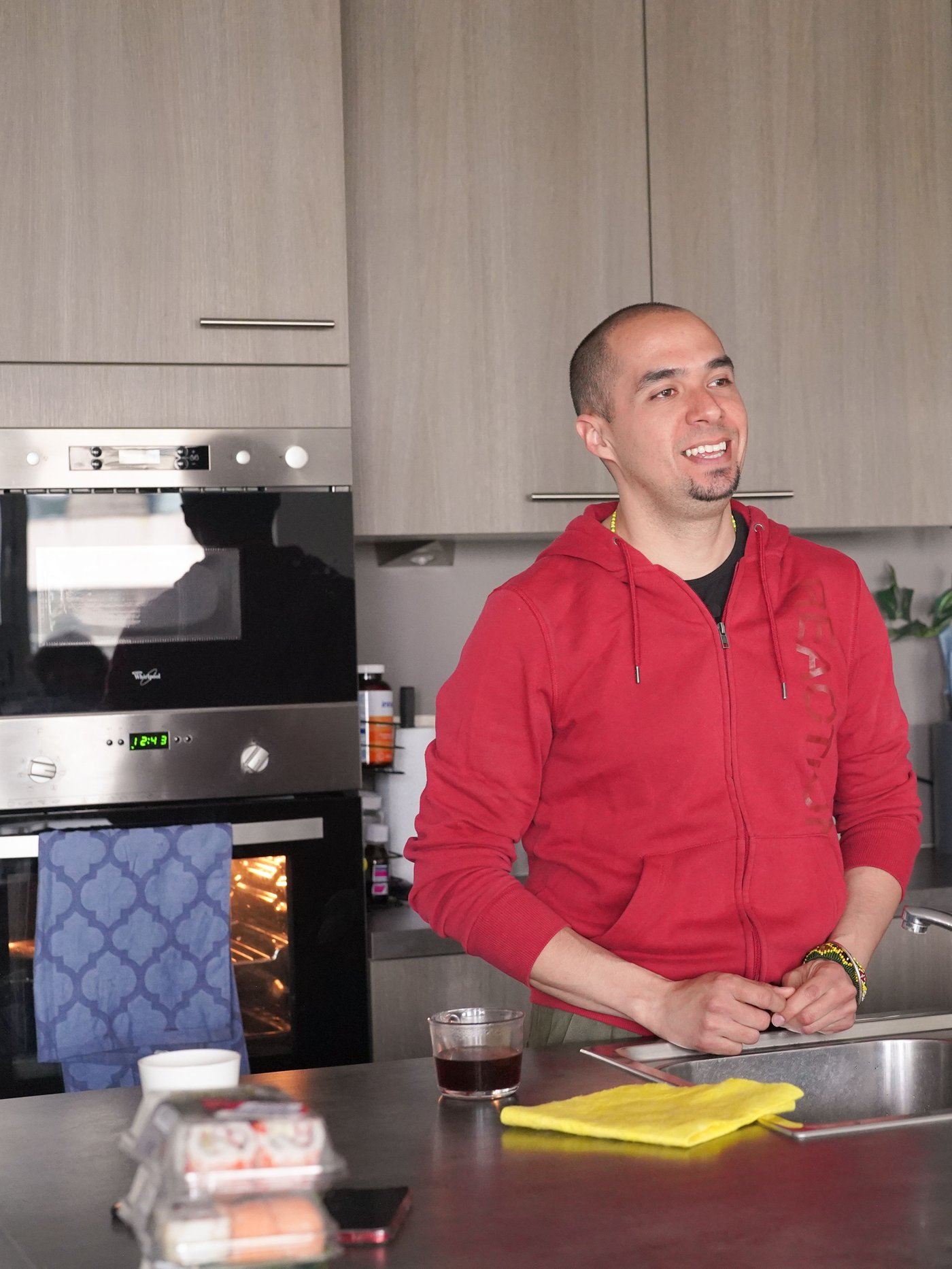  A man in a red hoodie standing in his kitchen smiling and drinking coffee. 