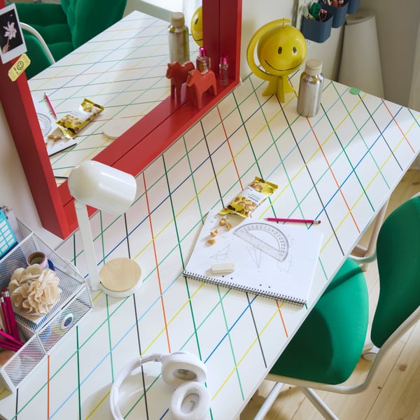 A LAGKAPTEN/SPÄND desk with a colourful geometric grid pattern paired with a green desk chair, white lamp and red mirror.