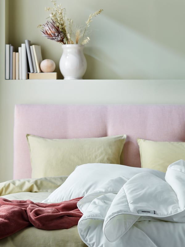 Una camera da letto con un letto con testata imbottita rosa e sullo sfondo un vaso e alcuni libri appoggiati ad una mensola. Il letto ha biancheria SMÅSPORRE.