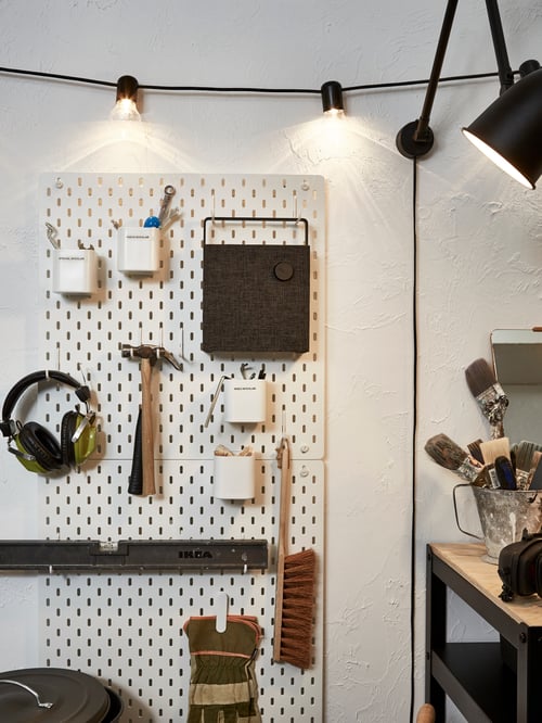 Un mur dans un garage avec deux panneaux perforés SKÅDIS blancs accueillant des outils et un haut-parleur accroché à un crochet sous une guirlande lumineuse près d’un plan de travail.