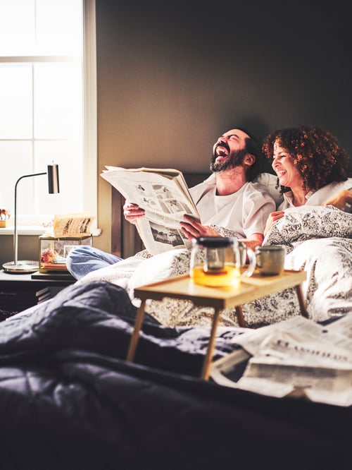Modern bedroom with two people laughing in bed, a bed tray, a work lamp, a duvet cover and two pillowcases.