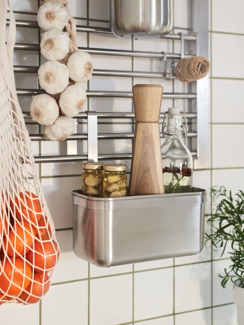 A kitchen wall with KUNGSFORS containers and hooks hanging on a wall grid, all in stainless steel, holding jars and garlic.