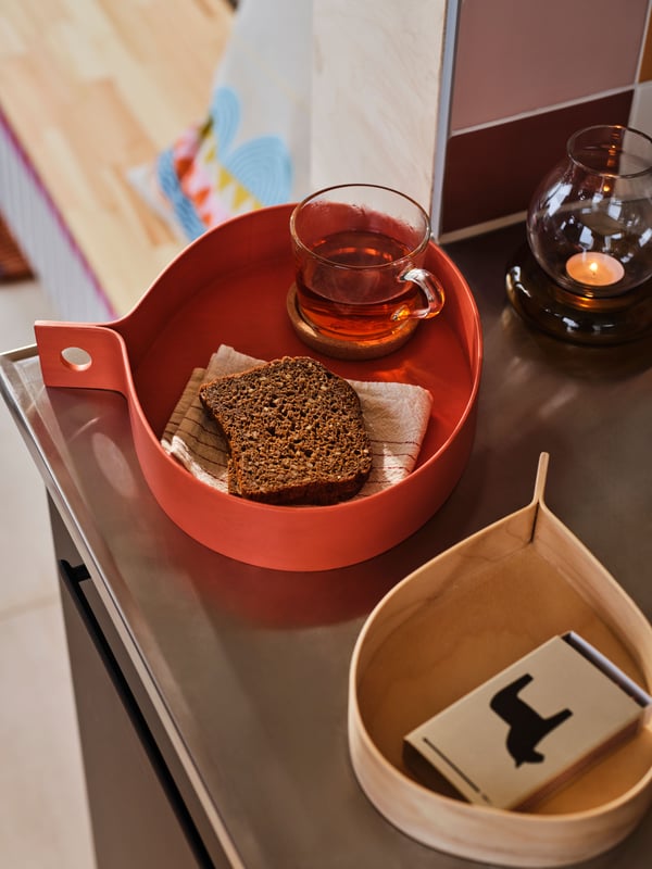  Una bandeja OMMJÄNGE roja y redonda sobre la que hay una rebanada de pan oscuro y una taza de cristal llena de té. A la derecha, hay una caja de cerillas HYLTA sobre una bandeja OMMJÄNGE en madera clara con forma de lágrima. En el fondo se encuentra una vela encendida en un portavelas.