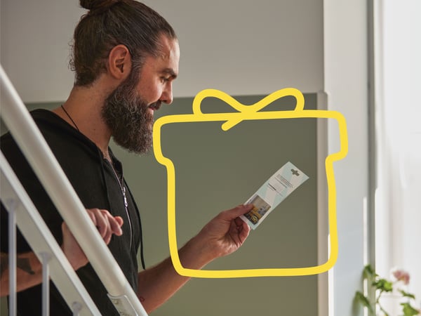 Man with a man bun standing by a white staircase at home, holding an IKEA gift card in his hand.