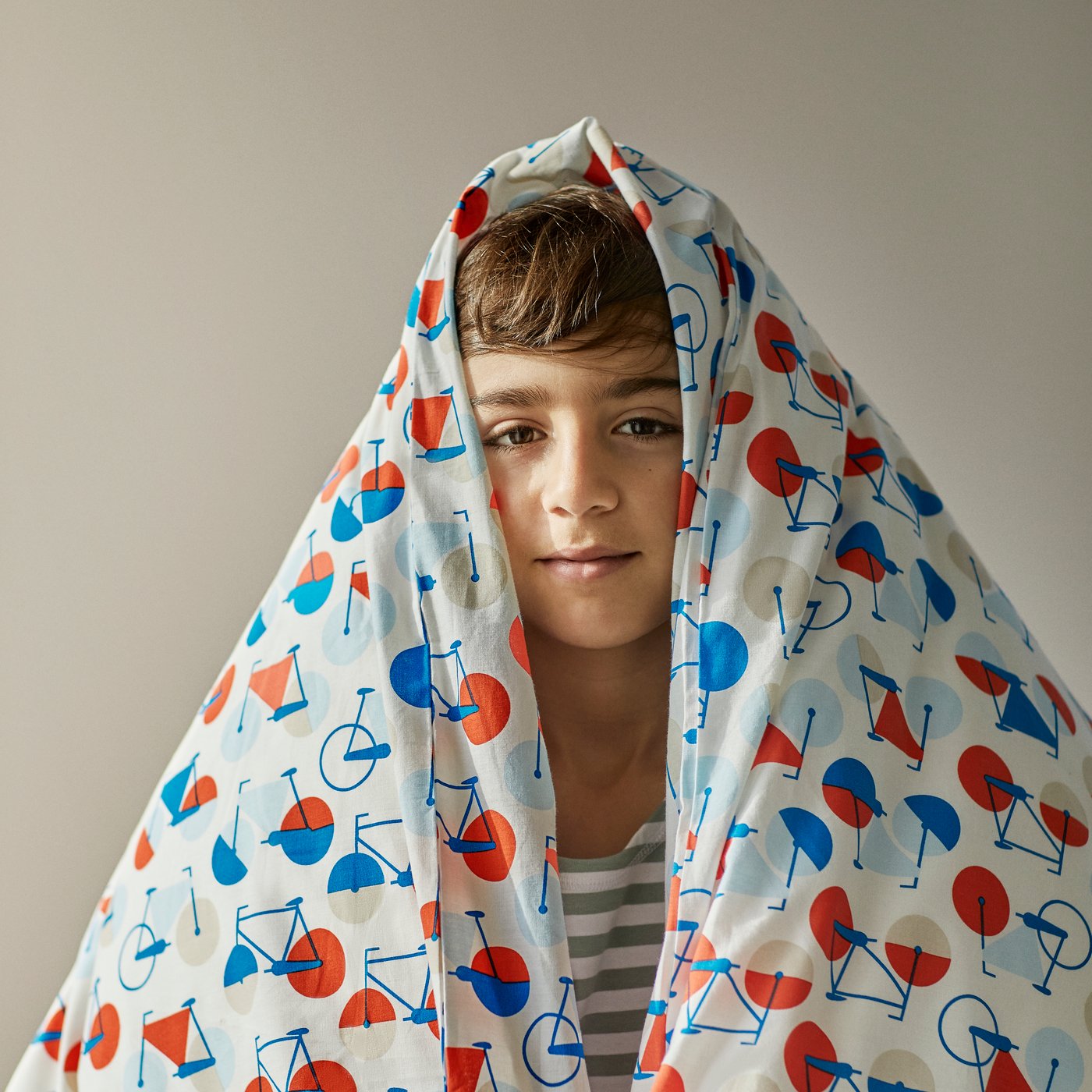 Un niño de unos 8 años con una sábana SPORTSLIG con dibujos de bicicleta en tonos azules y rojos sobre su cabeza.