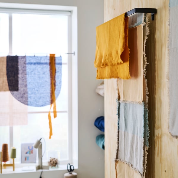 A black SKOGSVIKEN towel rail against a wooden wall holding different types of fabric near a desk with a FLOTTILJ lamp on it.