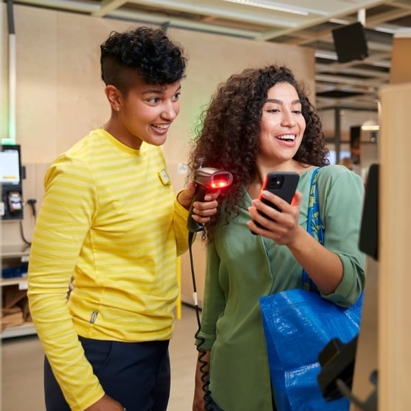 An IKEA co-worker smiling while scanning a customers smartphone at a self service check-out counter.