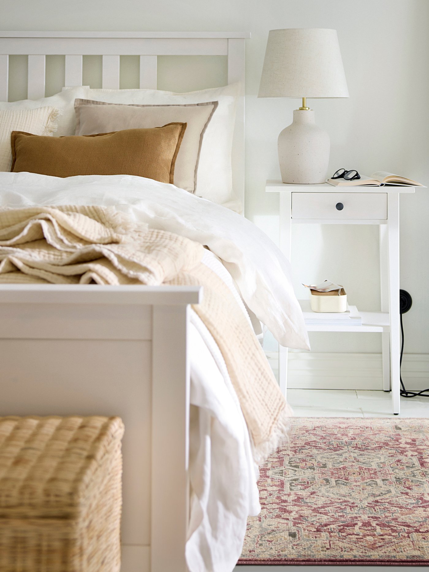 Ein roter JÄRNVÄG Läufer mit orientalischem Muster liegt neben einem weißen HEMNES Bettgestell. Auf dem Bett befinden sich weiße Bettwäsche, ein HUMLEMOTT Plaid in elfenbeinweiß und verschiedene Dekokissen in weiß und beige Tönen.