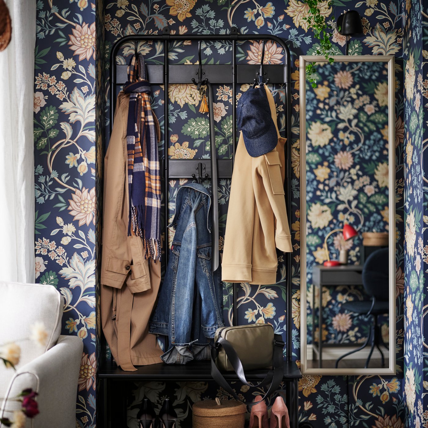 An einer Wand mit geblümter Tapete ist ein NIPÅSEN Garderobenständer mit Bank/Schuhaufbewahrung zu sehen, daneben befindet sich ein NYPONBUSKE Spiegel. Am Garderobenständer hängt Straßenkleidung.