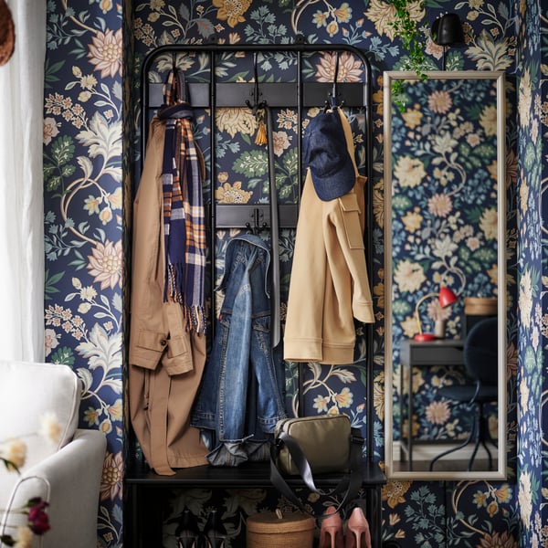 A NIPÅSEN coat rack and bench with shoe storage holding outdoor wear against a flowery wall with a NYPONBUSKE mirror by it.