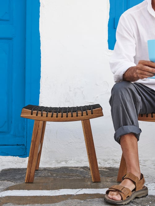 A person sits on a light brown, stained acacia STACKHOLMEN stool with a black braided rope seat by an identical, empty stool.