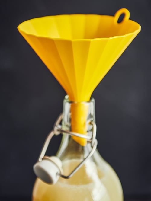 Vor einem dunklen Hintergrund wird eine KORKEN Flasche mit Verschluss mittels eines UPPFYLLD Trichters mit einer gelben Flüssigkeit befüllt.