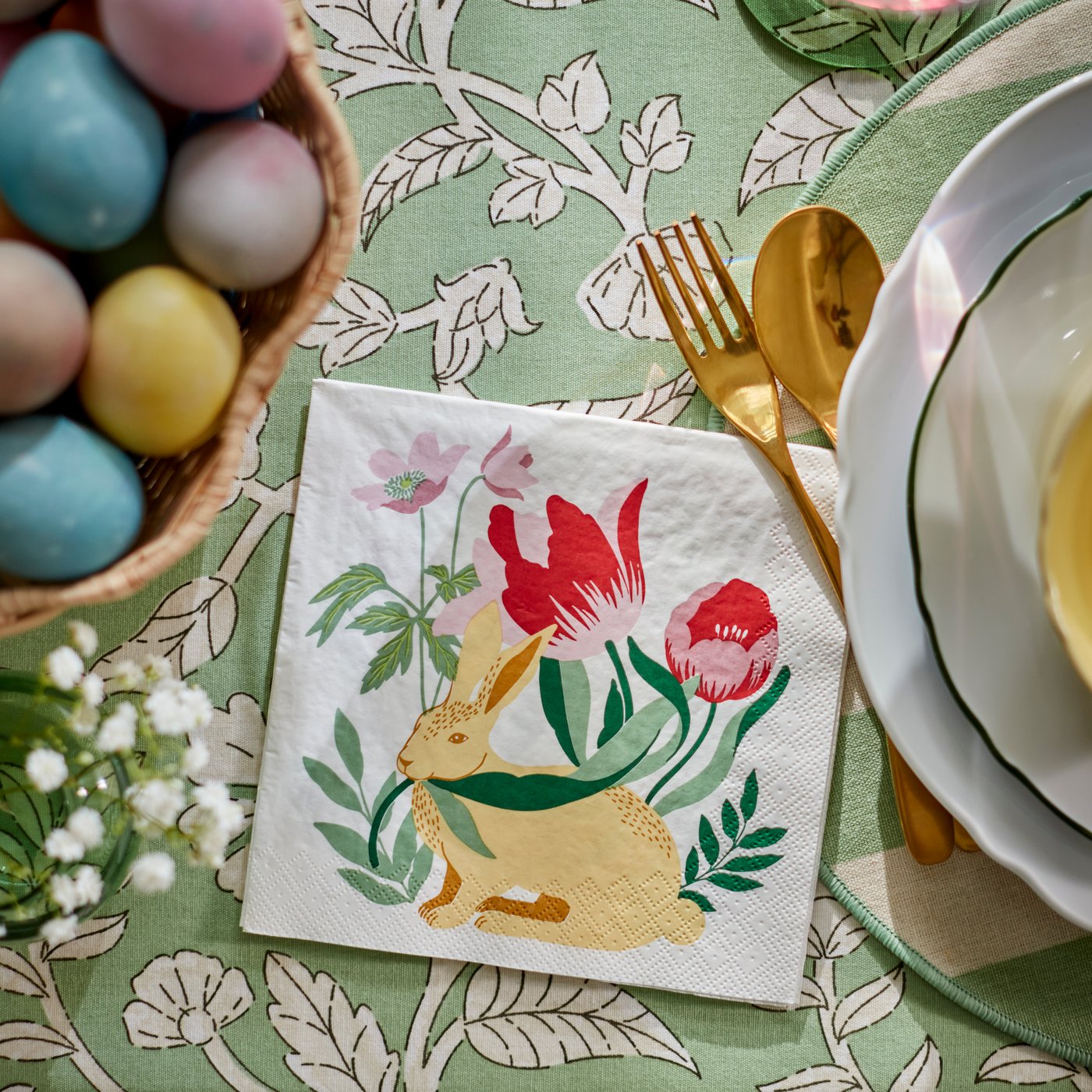 A close-up of an Easter bunny-patterned TJÄRLEK paper napkin, next to an UPPLAGA plate on a green SMÖRFISK tablecloth.