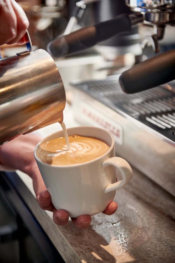Person häller skummad mjölk i kaffekopp och gör latte art, espressomaskin syns i bakgrunden.