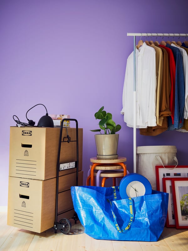 Moving-in-mess with DUNDERGUBBE moving boxes, blue carrier bags, stackable stools, a clothes rack with lots of shirts.
