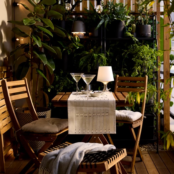 A plastic LAMPAN LED portable lamp with a white shade and clear base is lit, on the table of a bistro set in a lush balcony.