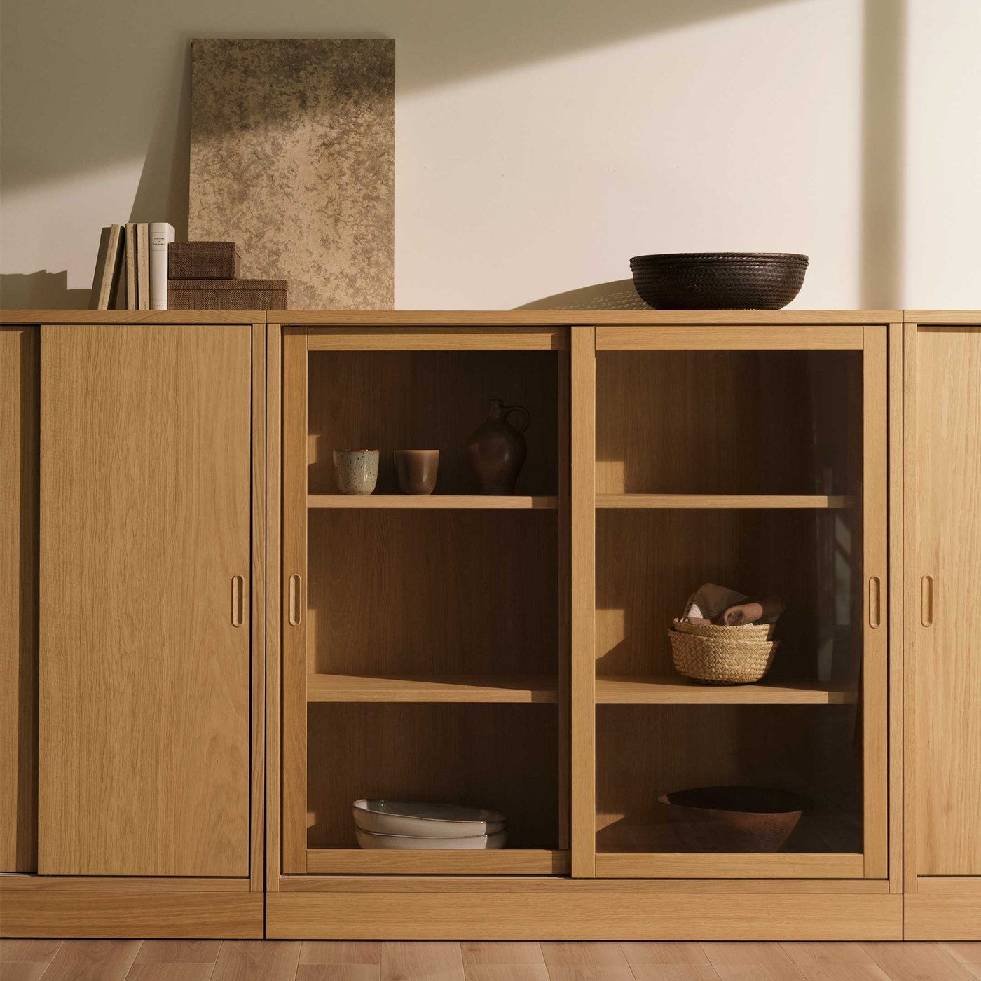 A beautifully organised wooden storage unit, perfect for keeping your living space tidy and stylish. This versatile piece combines closed cabinets for discreet storage with glass-fronted sections, allowing you to display your favourite items. Inside the glass sections, you'll spot neatly stacked plates, a charming woven basket, and some lovely ceramic cups and a vase, all ready to be used or admired. On top, a decorative dark woven bowl adds a touch of texture, alongside some books and an abstract art piece, creating a personal and inviting feel. The warm wooden tones and clean lines make this a wonderful addition to any home, helping you create a space that’s both functional and full of character.