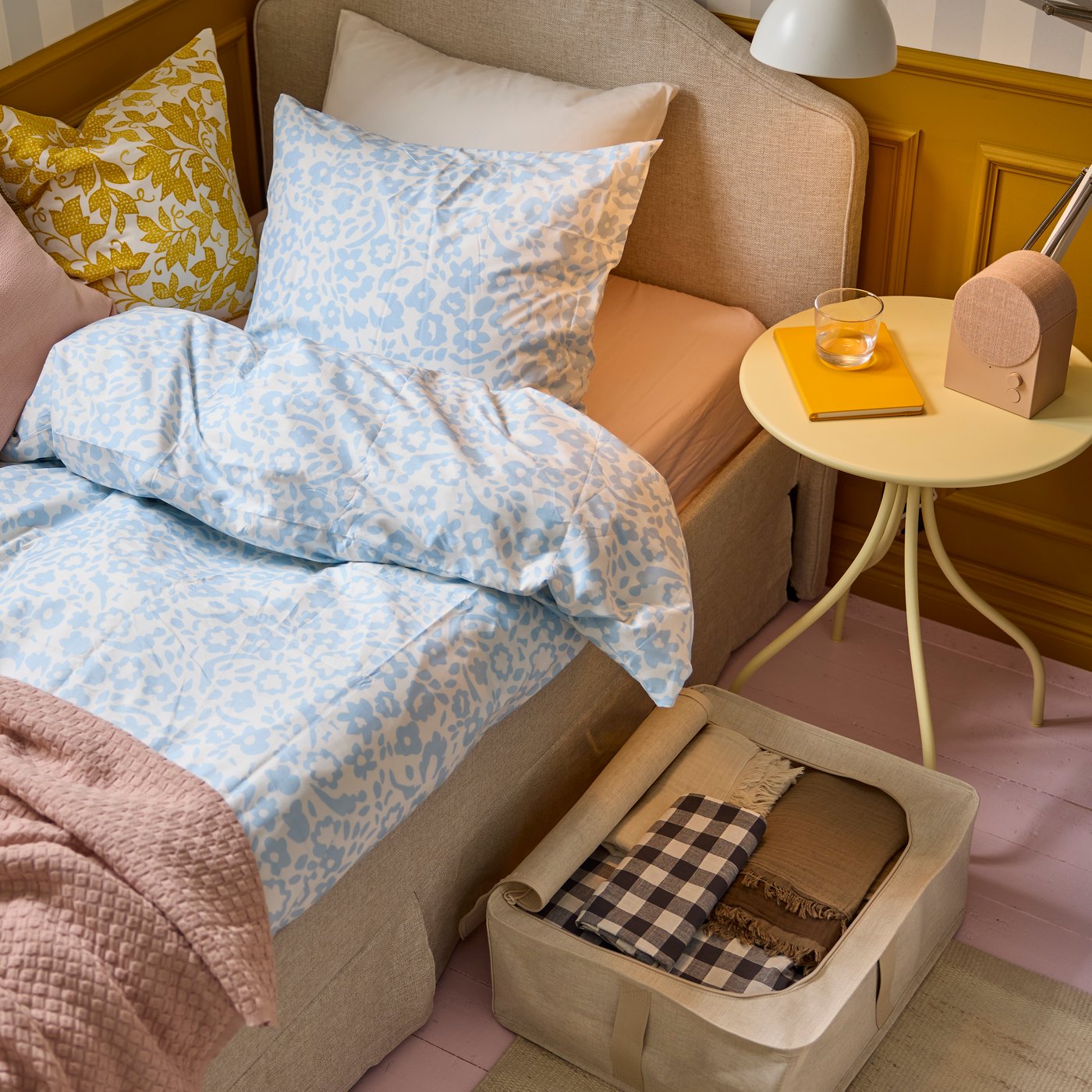 En bäddad säng med blåmönstrat påslakan och rosa överkast bredvid ett gult sidobord med glas och bok. Under sängen står en tygförvaringslåda med vikta textilier.