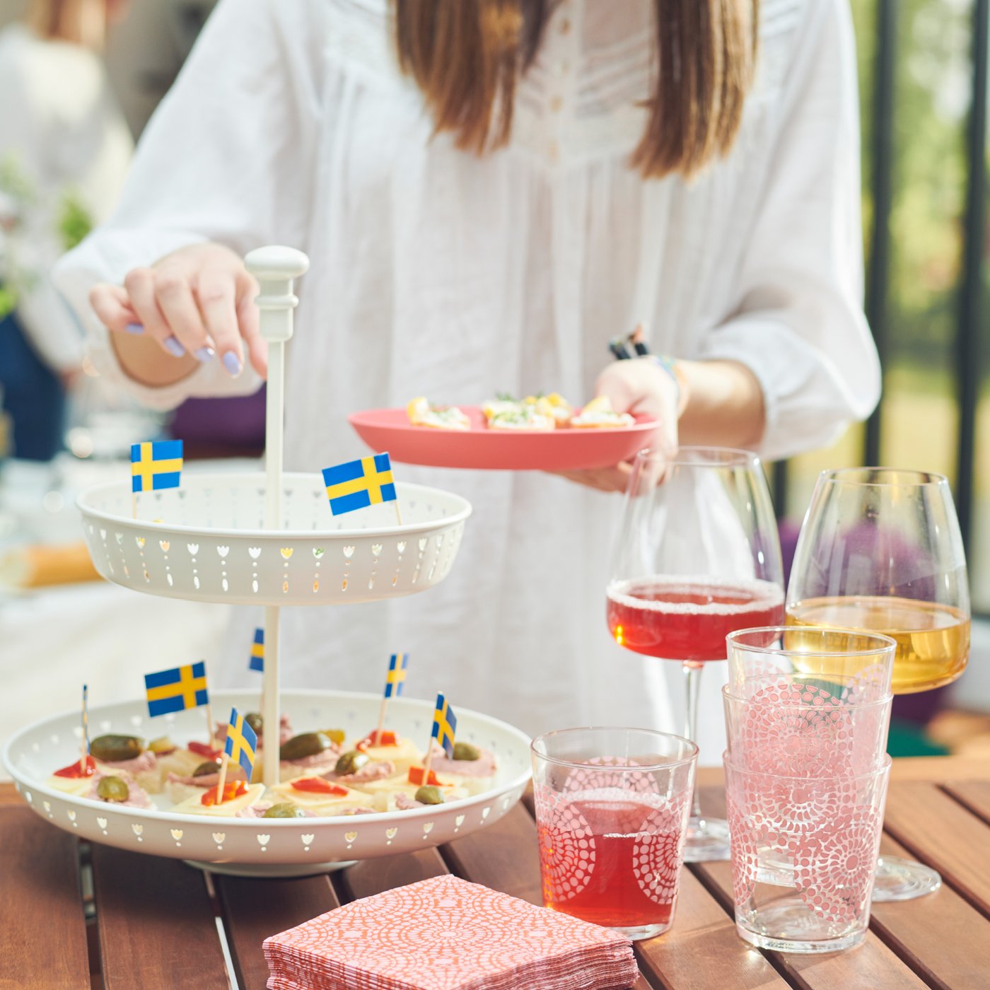 A person taking food from an IKEA serving stand on a wooden table, glasses and pink napkins on the side.