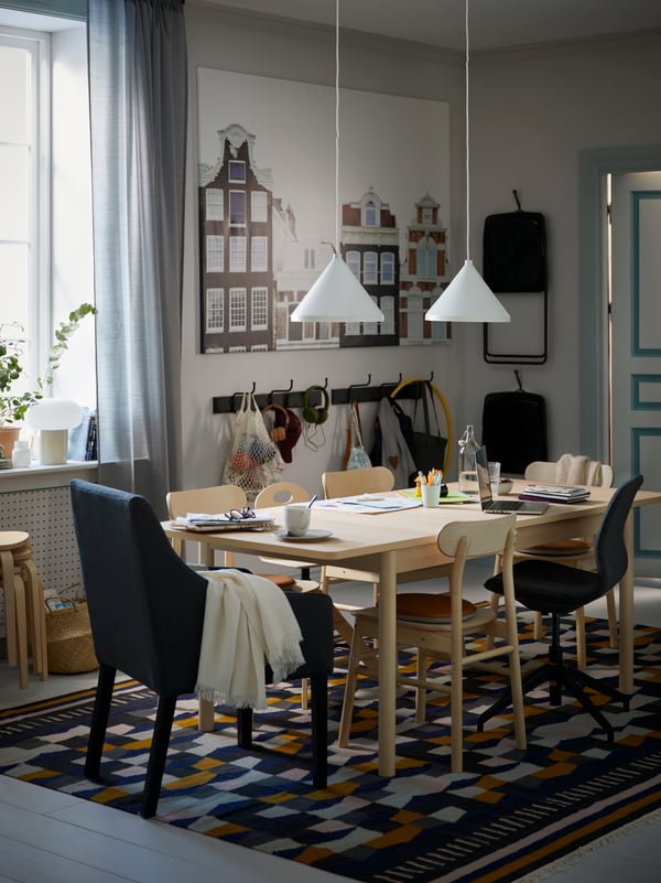 Una zona de comedor con sillas y una mesa de abedul RÖNNINGE sobre una alfombra TÅRBÄK multicolor, con lámparas de techo blancas NÄVLINGE encima.