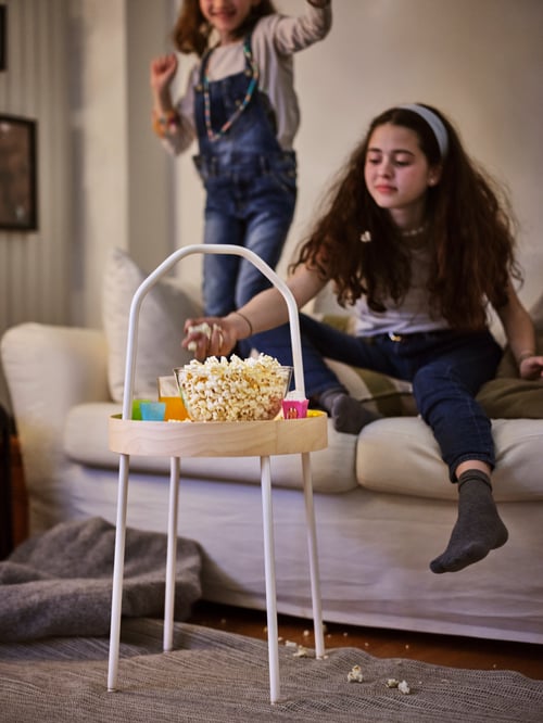 Zdjęcie dwóch małych dziewczynek siedzących na sofie, z których jedna chwyta garść popcornu z przezroczystej miseczki stojącej na okrągłym stoliku bocznym. 
