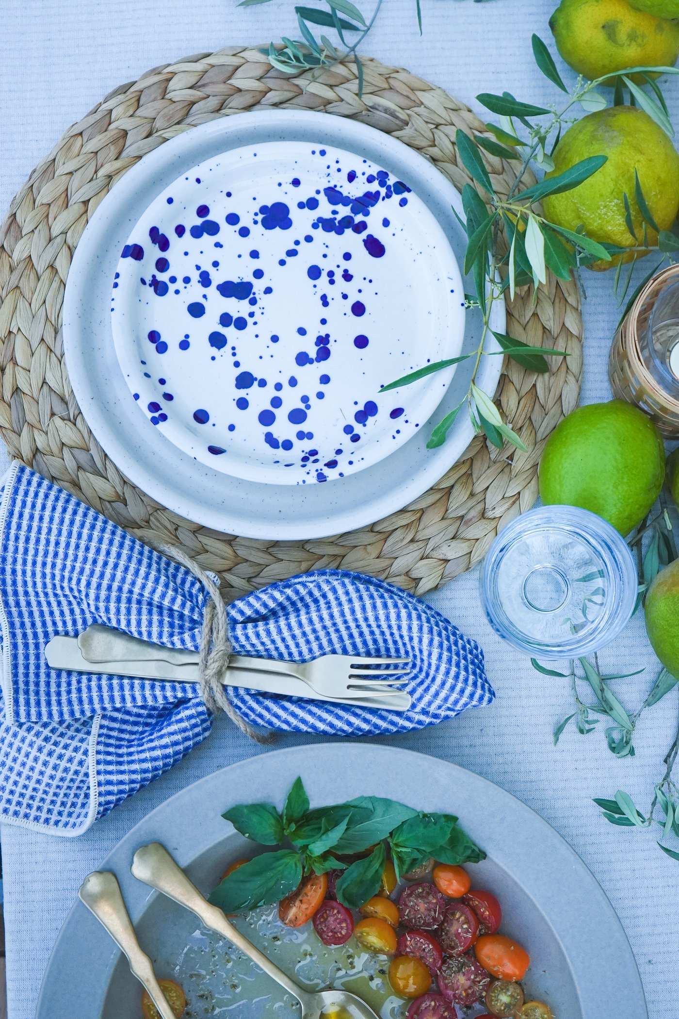 Décoration de table méditerranéenne avec de la vaisselle SILVERSIDA bleue et blanche posée sur des sets de table en raphia, des couverts enroulés dans une serviette à carreaux, des citrons verts frais et une salade de tomates cerises au basilic.