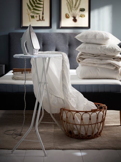 Bedroom area featuring a white ironing board with a clothes iron and a sheet that's partially inside a basket.