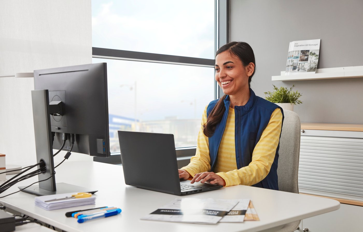 A smiling person in a yellow sweater and a blue vest is working on a black laptop placed on a white desk next to a window.