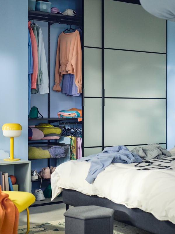 Lit matelassé SLATTUM gris foncé près d’une armoire-penderie intégrée SKYTTA/BOAXEL avec portes coulissantes dont une est ouverte.