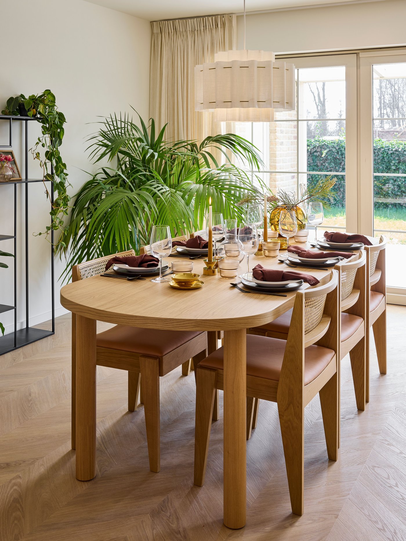 Ronde houten eettafel met stoelen, gedekte tafel en planten naast raam met gordijnen en tuinzicht.