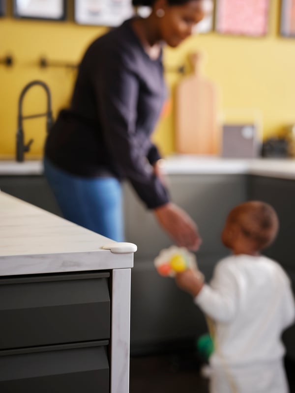 A woman and a toddler in a kitchen with a white UNDVIKA corner bumper on the corner of a white marble effect worktop.