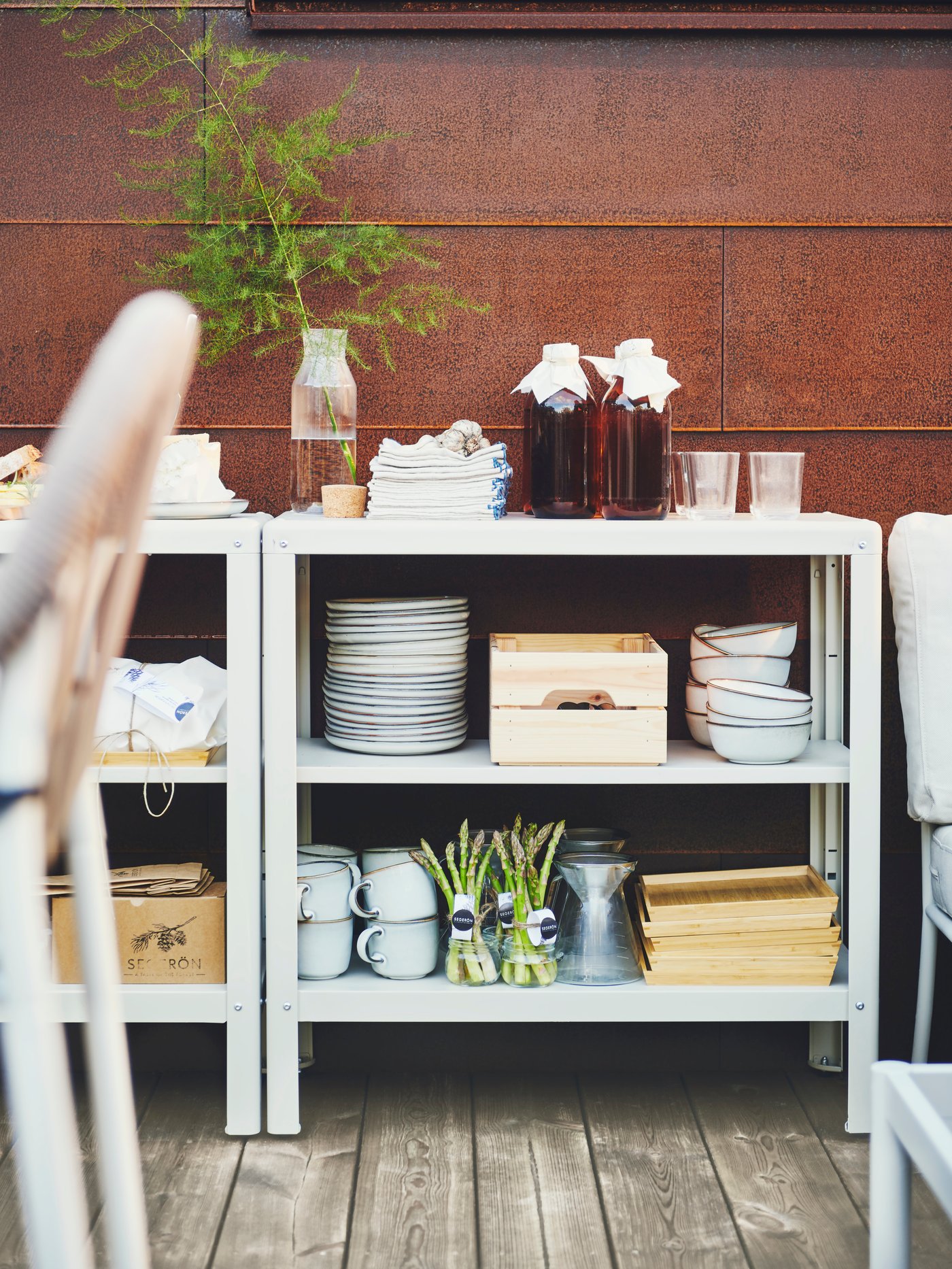 Espacio al aire libre con dos estanterías KOLBJÖRN con una pila de platos, cuencos, tazas, cajas y una cafetera.