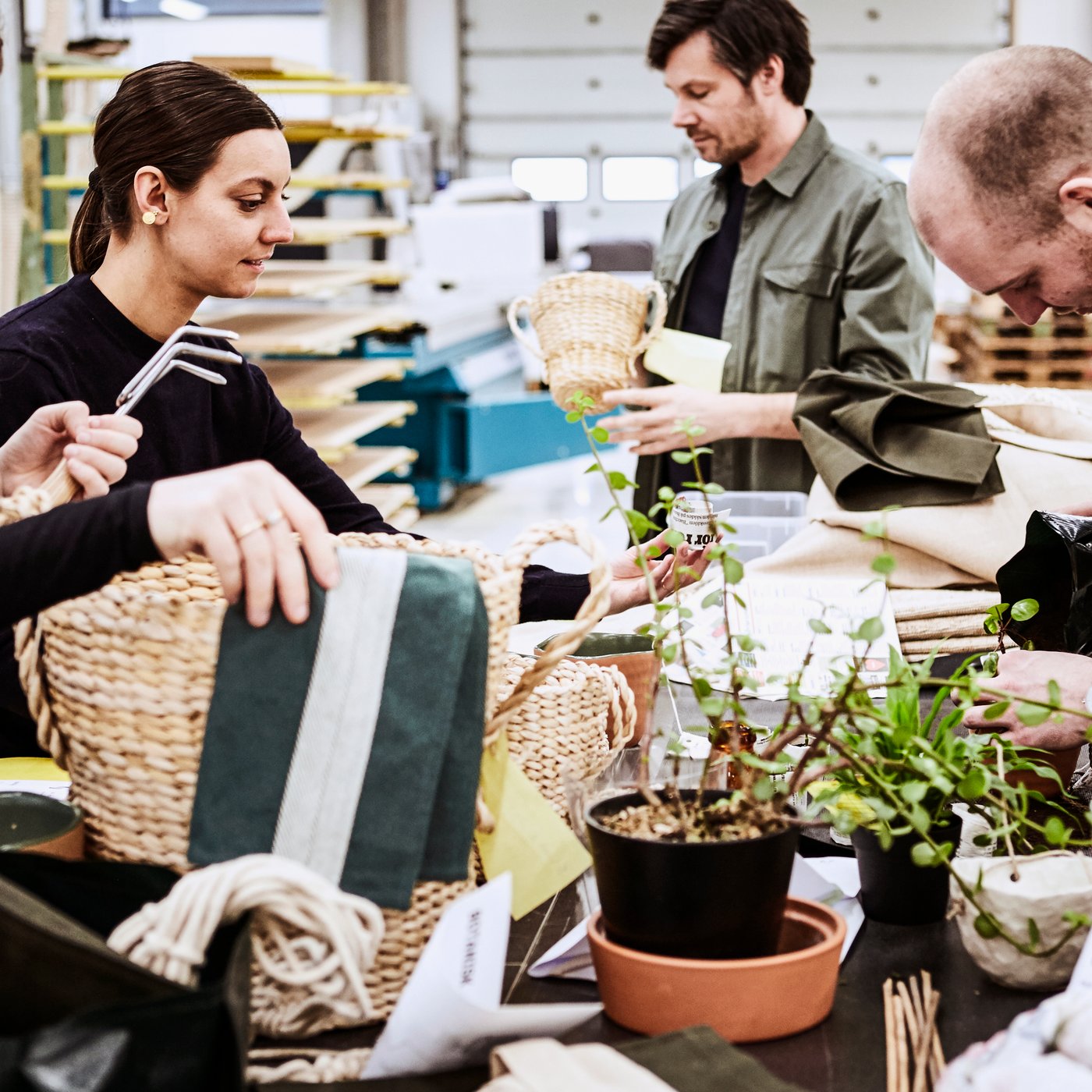 Four IKEA designers inspecting woven baskets around a large work top filled with bolts of folded material and pot plants.