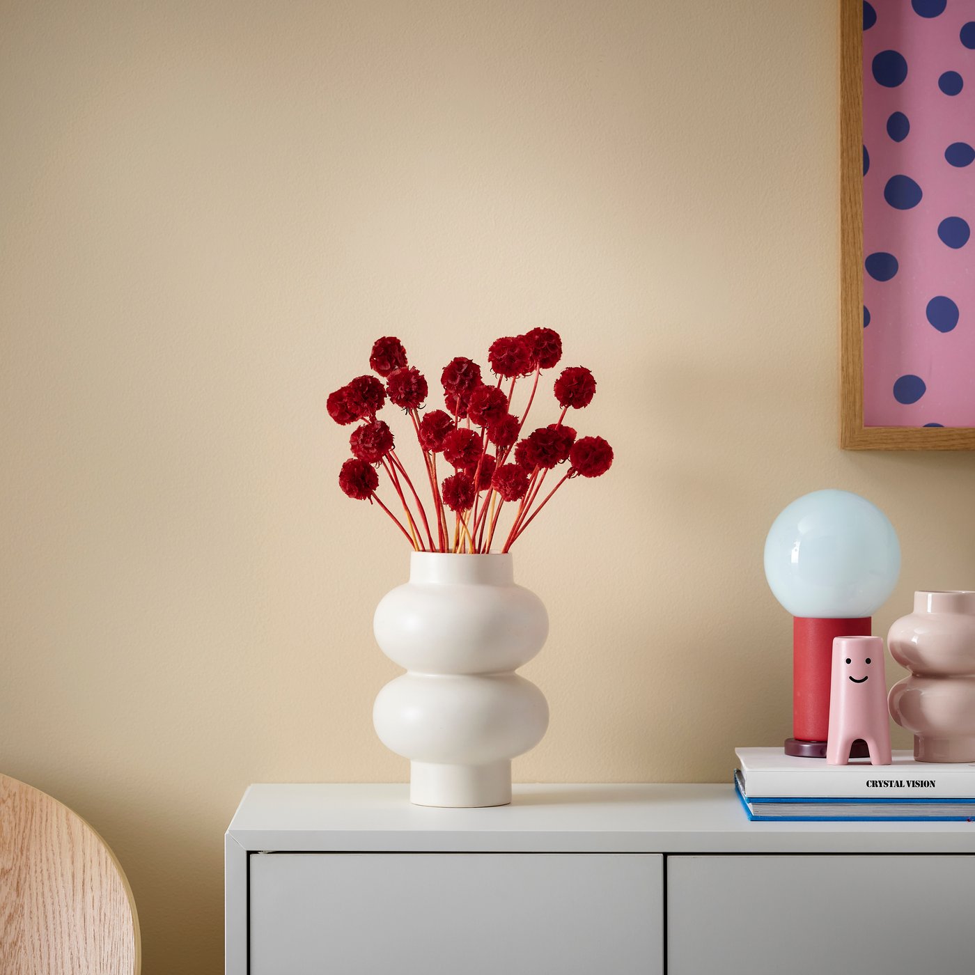 A beige vase with red dried flowers stands on a sideboard with colorful decorations next to it.