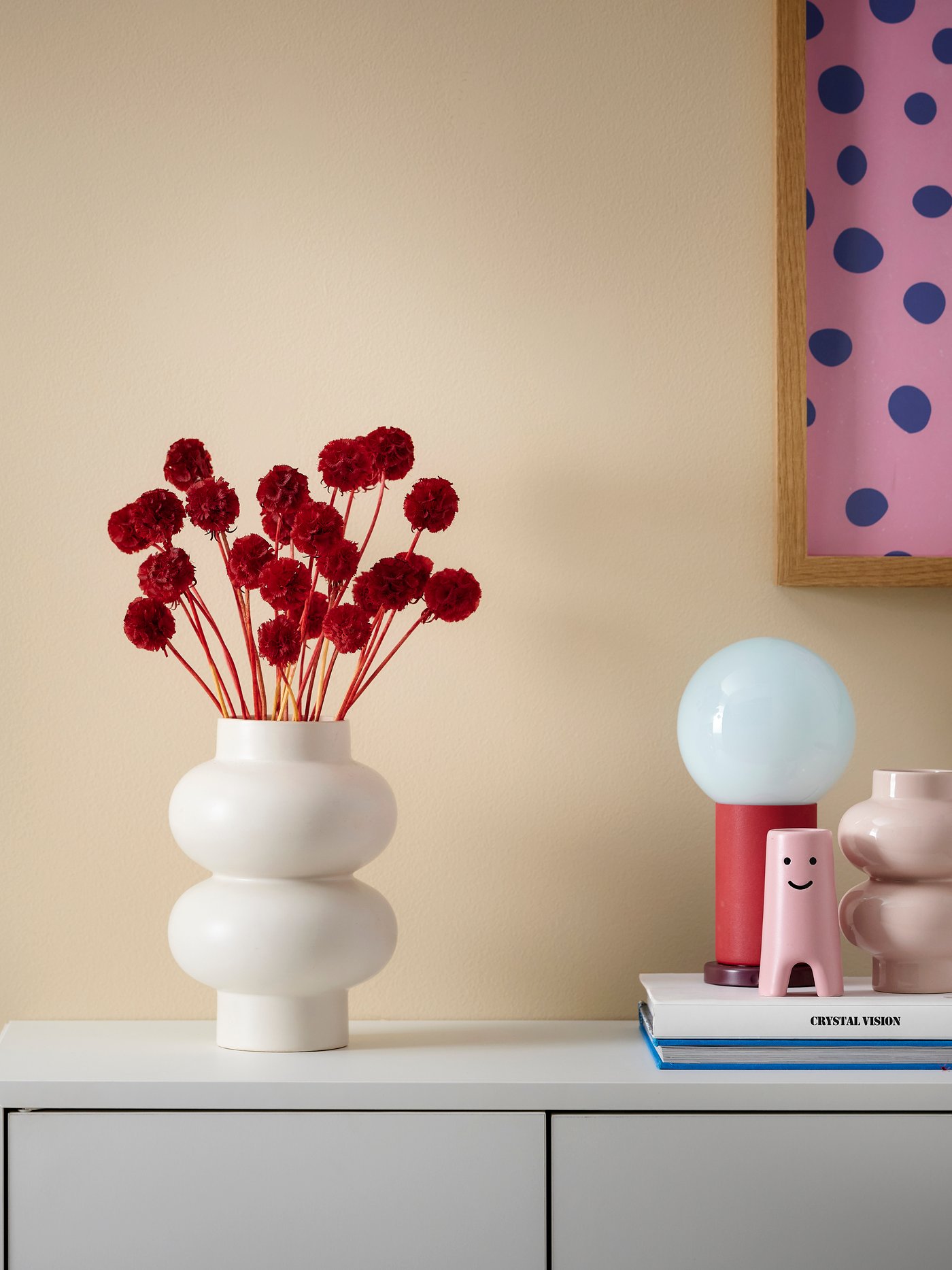 Auf einem Sideboard steht eine weiße Vase mit roten Trockenblumen und danaben eine rote Tischlampe und rosane Dekofiguren.