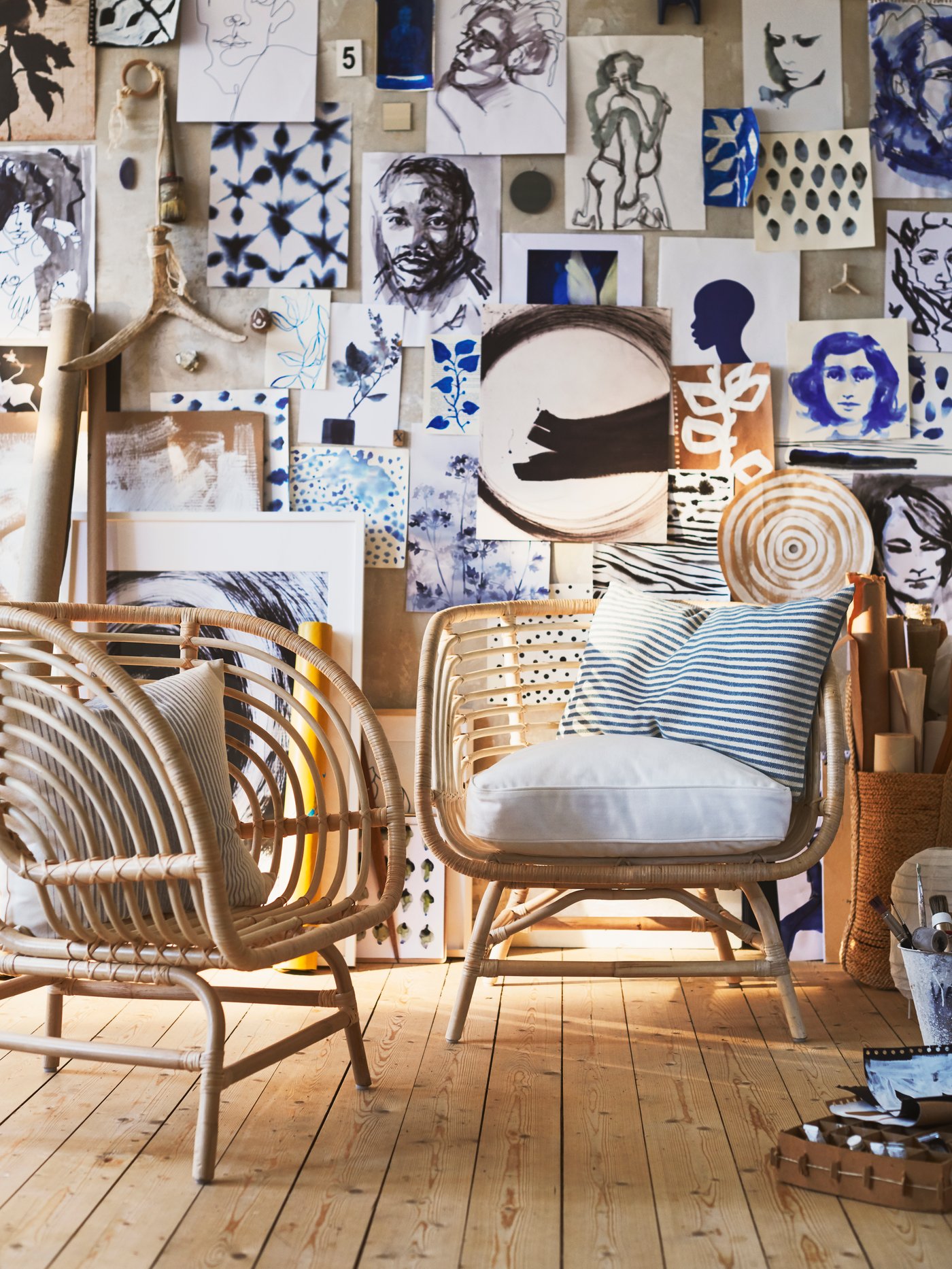 Två BUSKBO rottingfåtöljer med kuddar står på trägolv framför en vägg fylld med konstverk, teckningar och blåmönstrade bilder i olika storlekar.