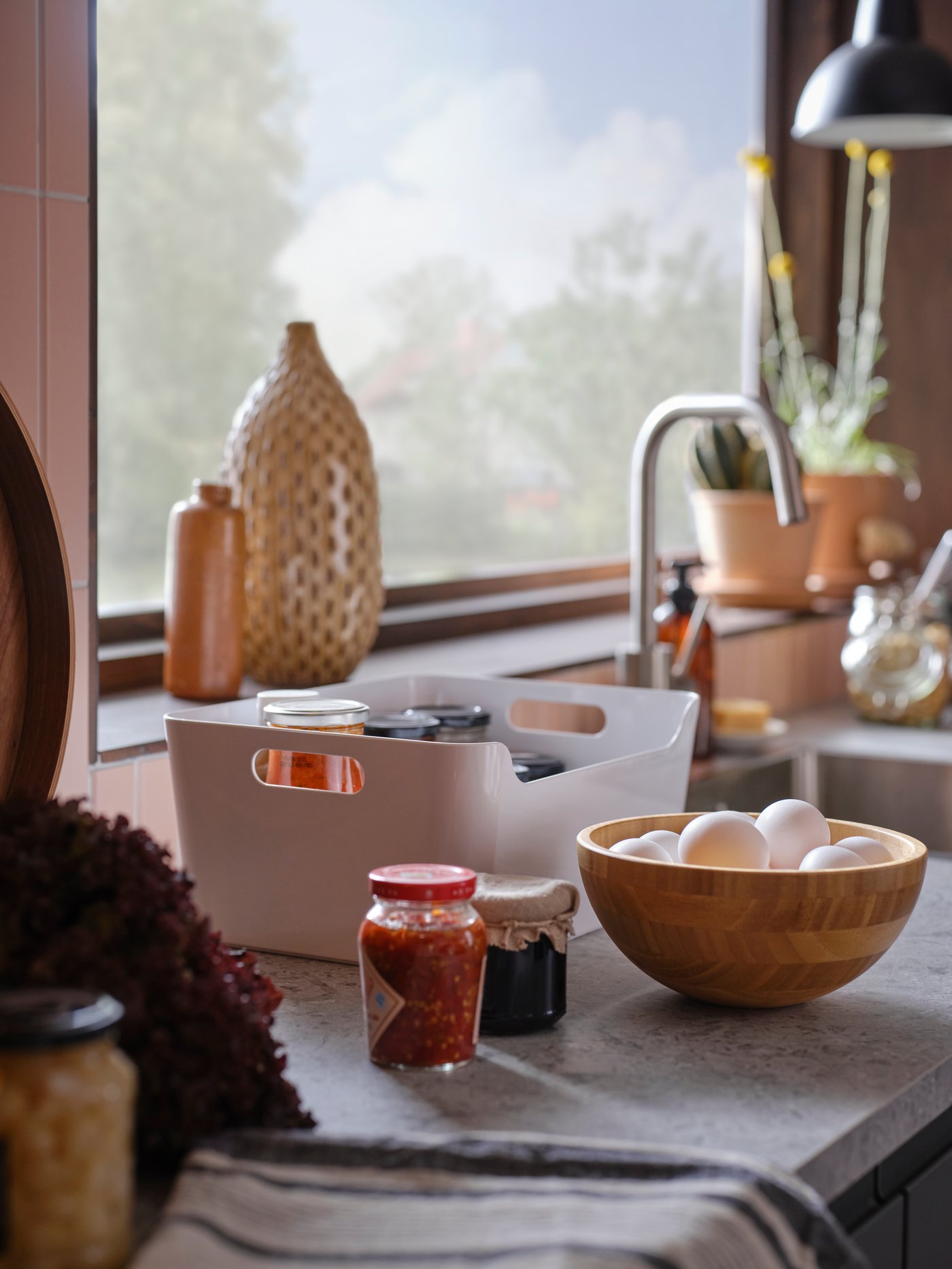 A white VARIERA box with jars in it next to two more jars and a bamboo bowl with eggs, on a kitchen worktop by a window.