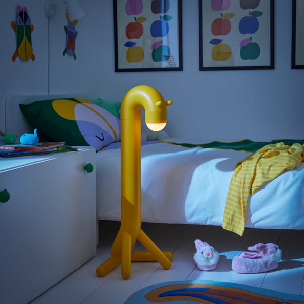 Yellow giraffe-shaped GREJSIMOJS floor lamp standing in a dark room beside a child’s bed, creating a gentle glow.