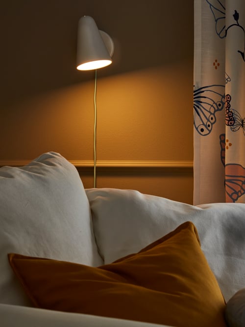 A white FUBBLA wall-lamp is lit and on a mustard-coloured wall by curtains and an armchair with white and yellow cushions.