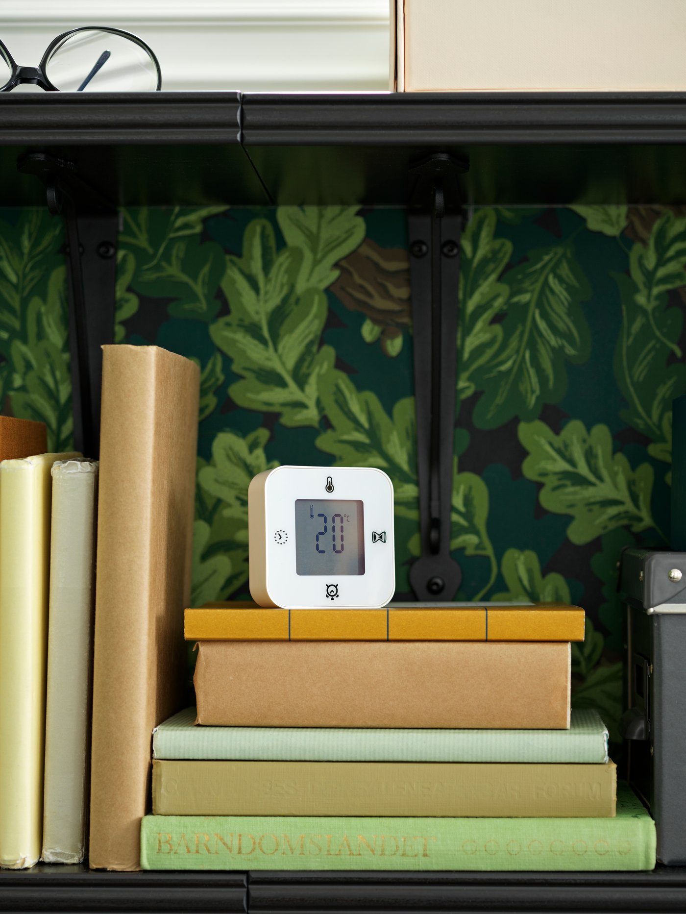 Egy KLOCKIS óra/hőmérő/ébresztő/időzítő áll könyvek tetején egy polcon, egy levélmintás tapétával borított fal előtt.